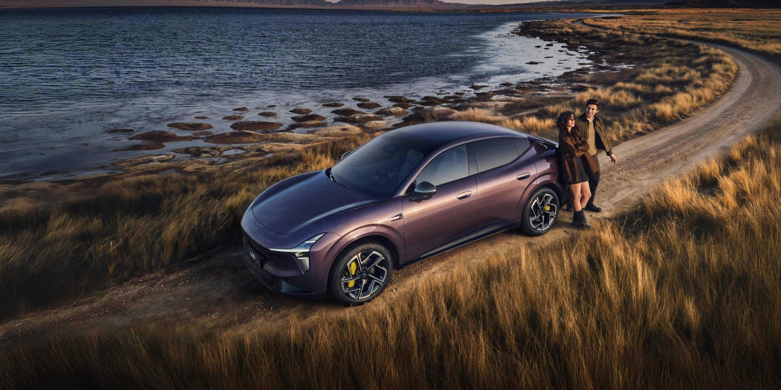 Xpeng L03 mauve gar&eacute; sur un chemin de terre au bord d'un lac, au coucher du soleil, avec un couple appuy&eacute; contre la carrosserie.