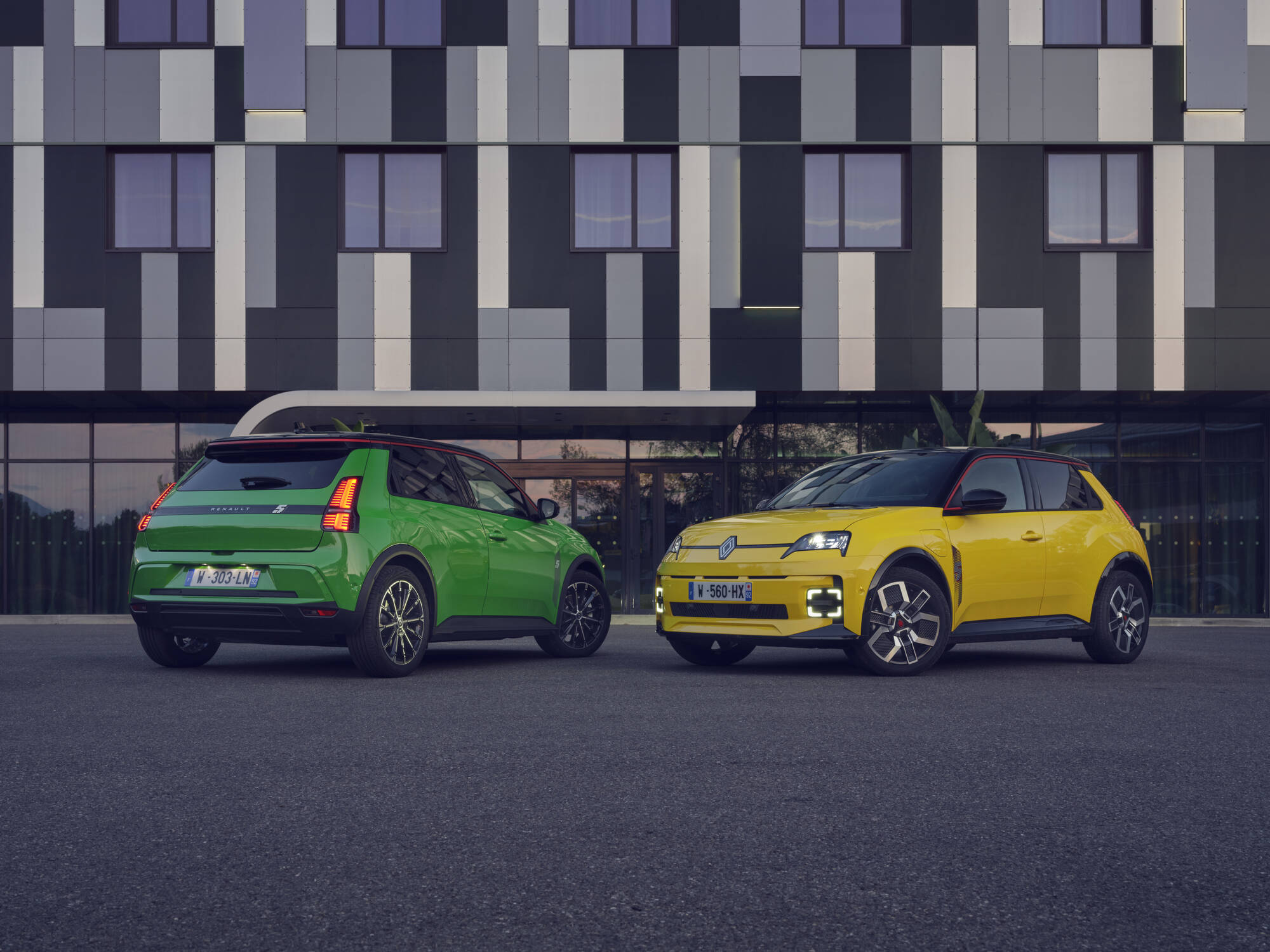 Deux Renault 5 E-Tech &eacute;lectriques, une verte et une jaune, gar&eacute;es devant un b&acirc;timent moderne.