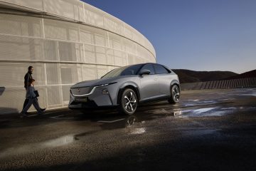 Mazda CX-6e gris, vue de trois quarts avant, devant une architecture moderne courbe